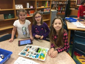 Three young students at a classroom table smile at the camera as they build with legos while reading directions from a tablet.