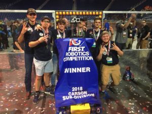 Six high school students smile at the camera while posing with their winning robot, which is draped in a winner's banner.